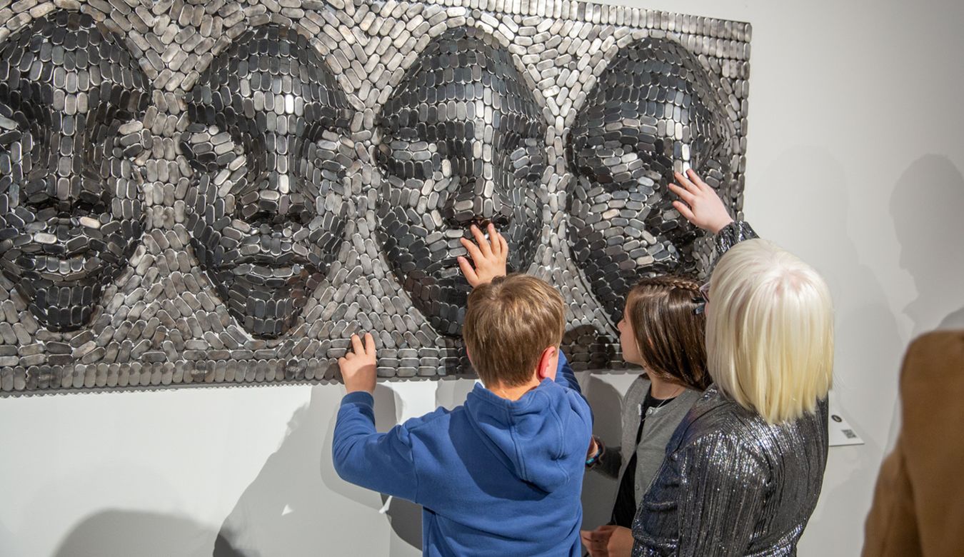 3 osebe se dotikajo skulpture.  Upodobljeni so štirje obrazi v mozaičnem slogu. Obrazi so izbočeni iz skulpture.