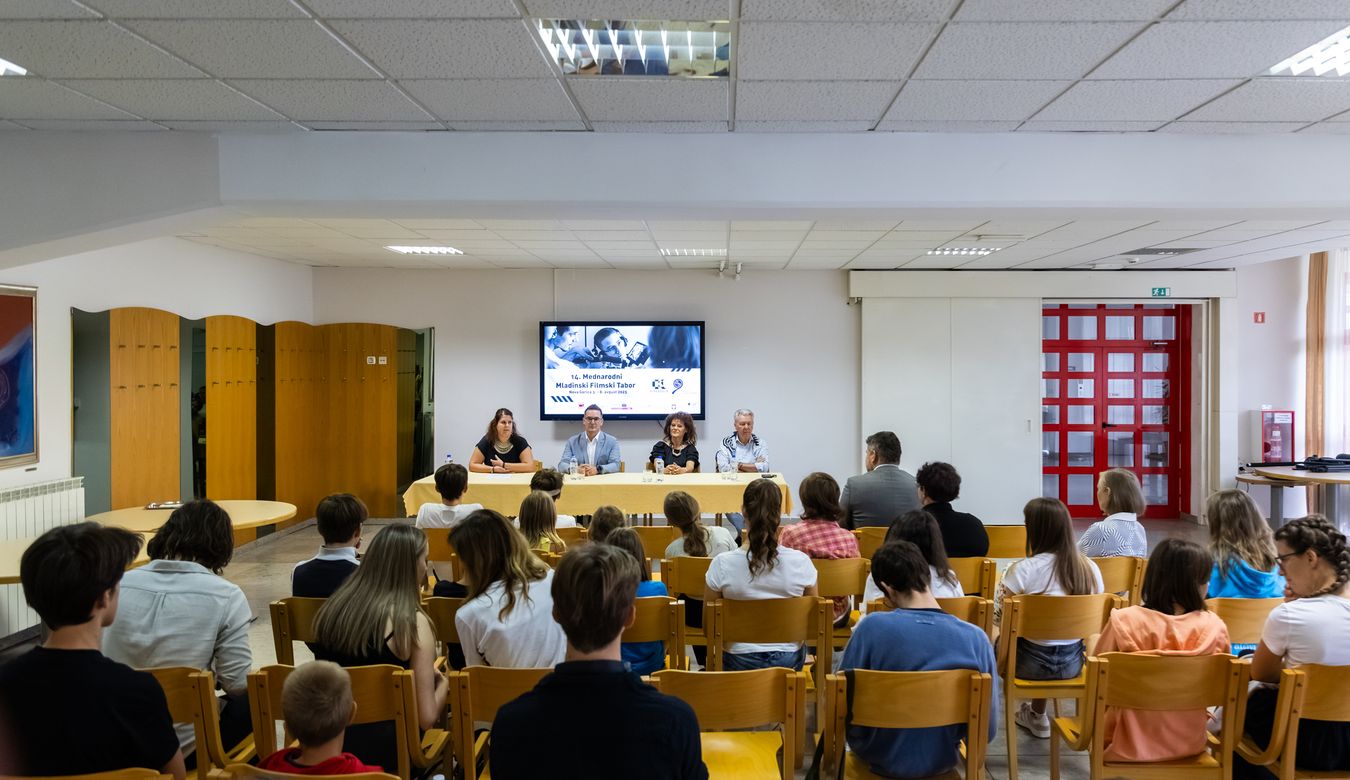 Fotografija pogleda proti govorcem, izza obiskovalcev novinarske konference.