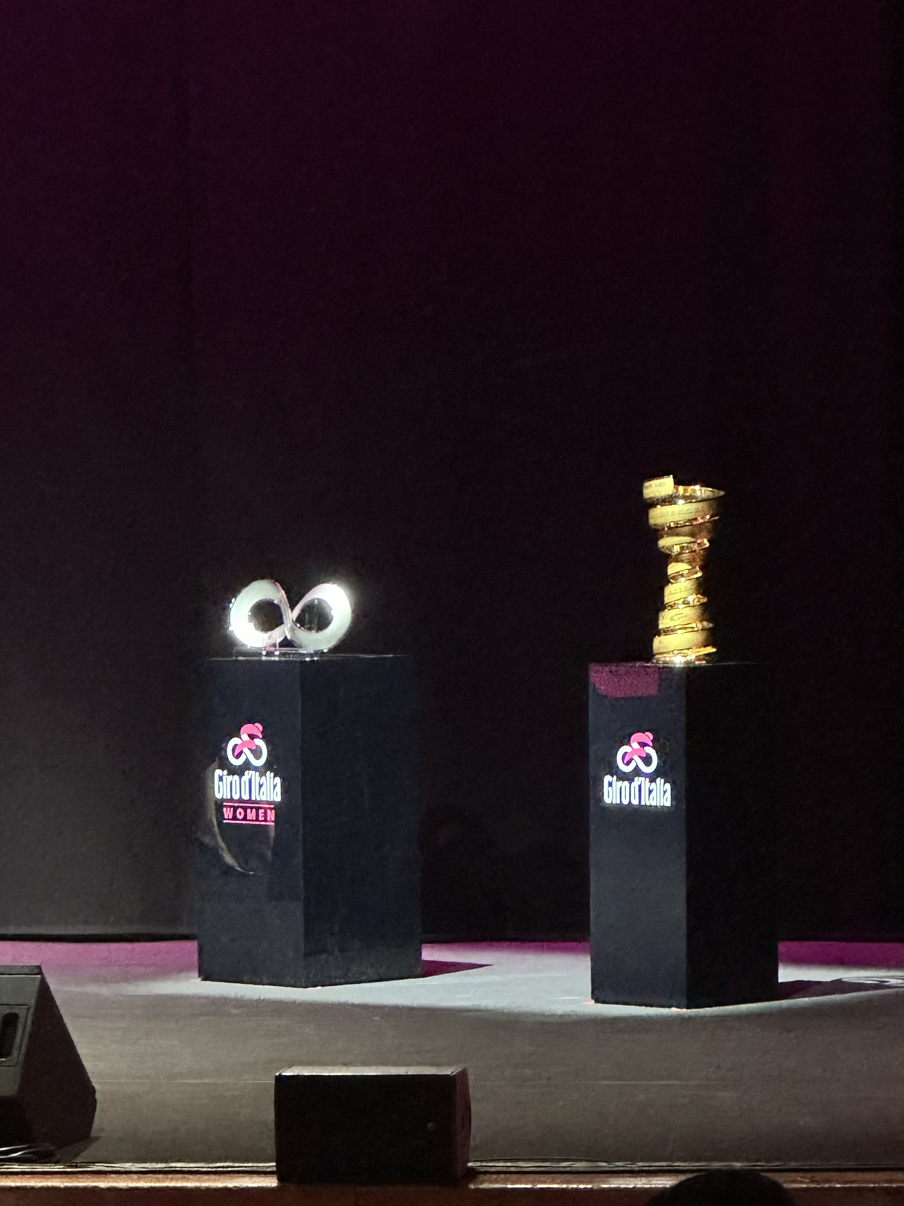 Na dveh črnih pravokotnih podstavkih sta postavljena trofeja Dirke po Italiji, zlat pokal in srebrn o priznanje, v obliki številke 8.