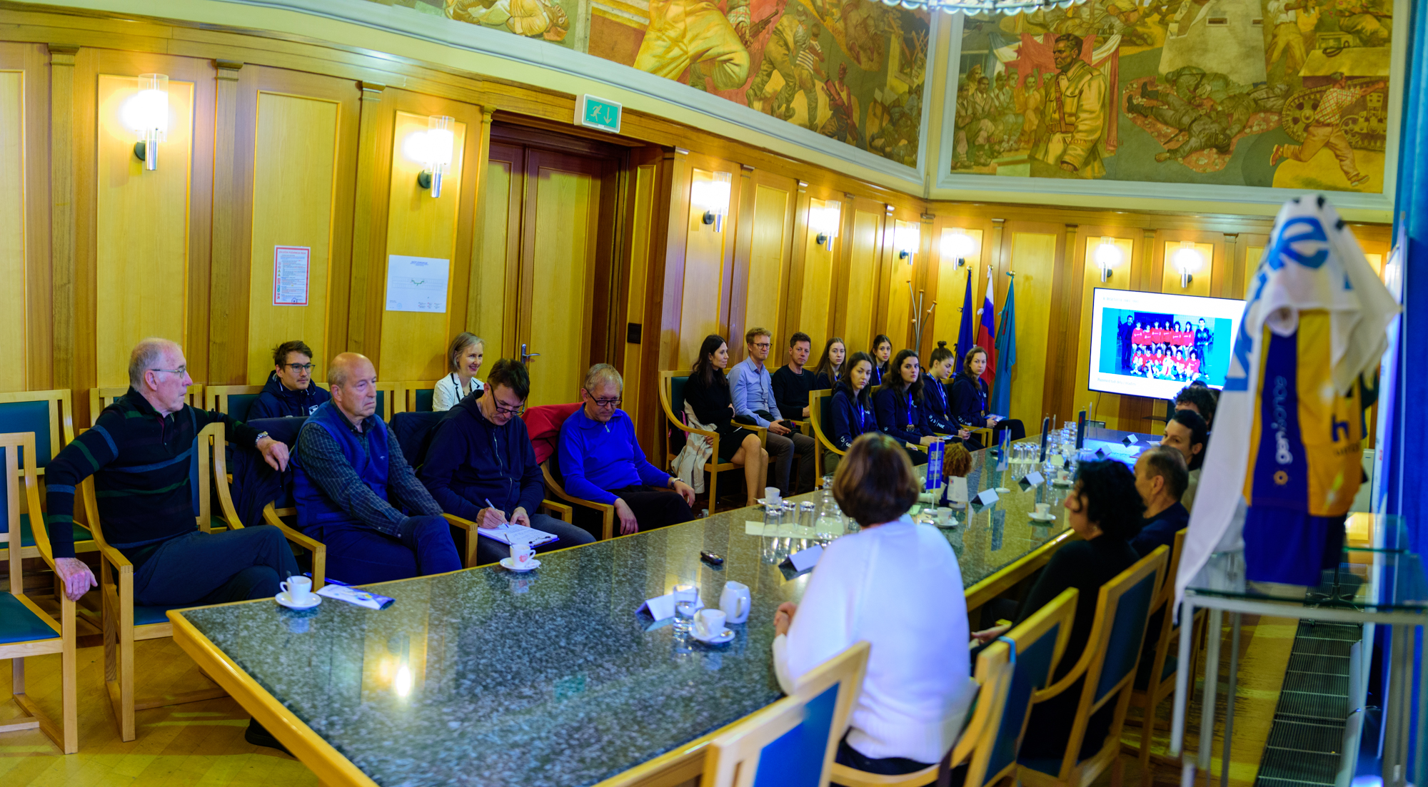 Udeleženci tiskovne konference sedijo za veliko mizo ali na stolih v Zeleni dvorani občinske stavbe