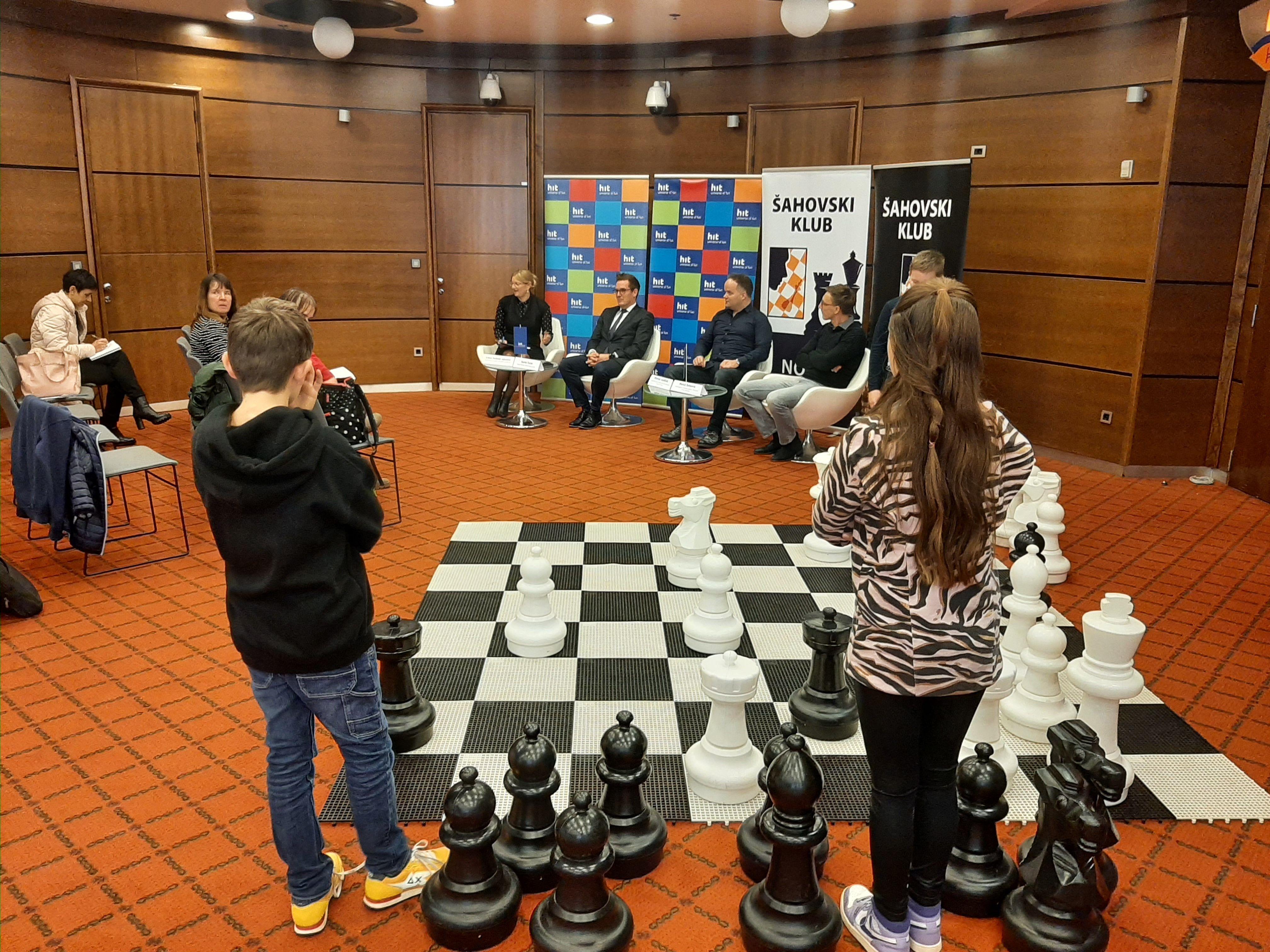 Fantek in deklica stojita ob večji črno-beli šahovnici s črno-belimi šahovskimi figurami