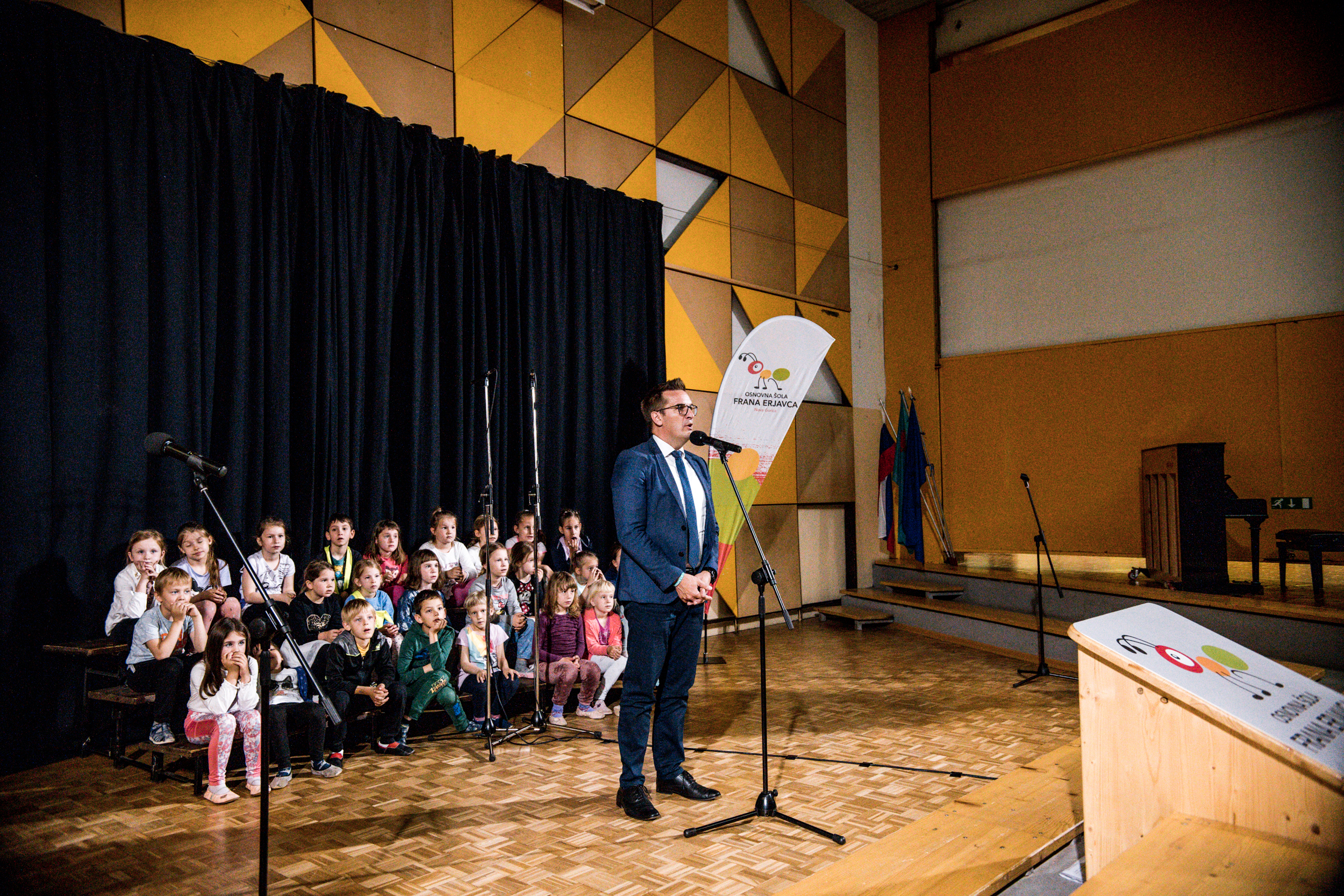 Novogoriški župan nagovarja občinstvo na OŠ Frana Erjavca, otroci sedijo v ozadju